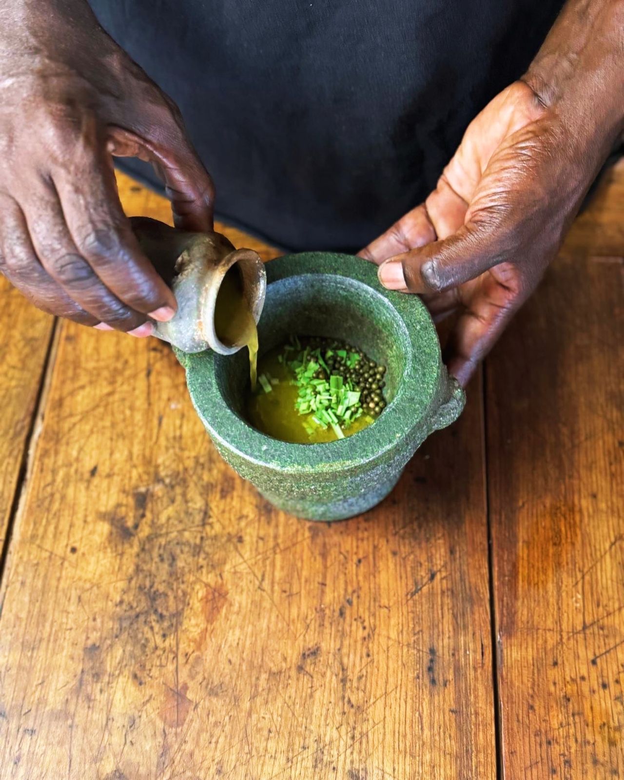 hands pouring oil into a green stone mortar of chopped herbs and peppercorns